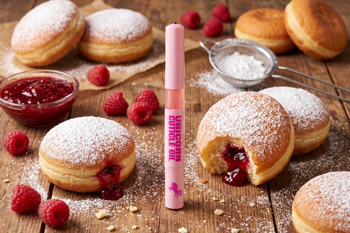 A pink tube labeled 'UNICORN OIL' stands upright on a rustic wooden table surrounded by powdered donuts, fresh raspberries, and a small bowl of red jam.