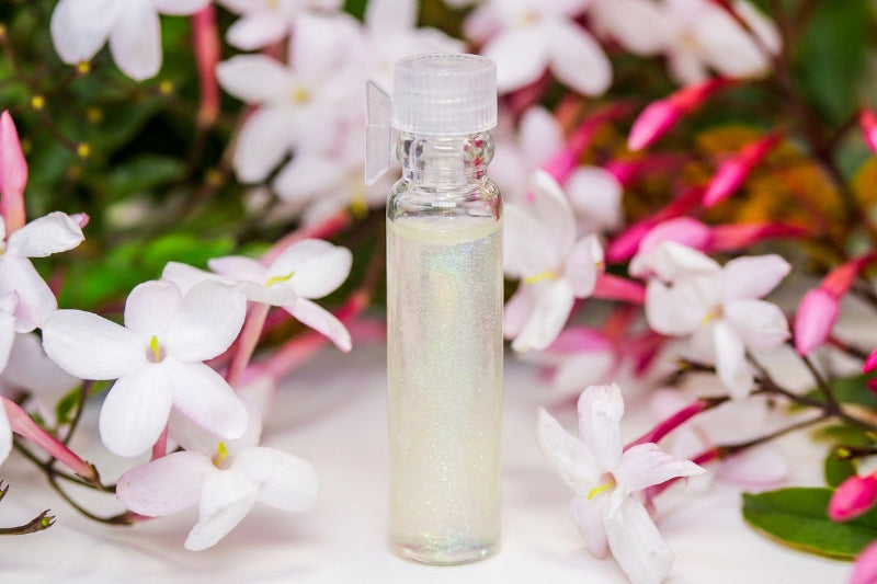 A small clear plastic bottle filled with pale yellow iridescent liquid, set against a background of white and pink flowers.