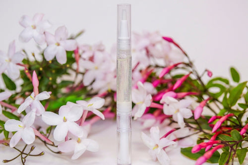 A clear cuticle oil pen standing upright in the center, surrounded by white and pink flowers with green leaves on a light background.