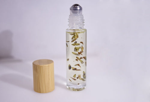 A clear glass roller bottle filled with a pale yellow liquid and containing dried botanical pieces, shown next to a light wood cap on a plain white background.