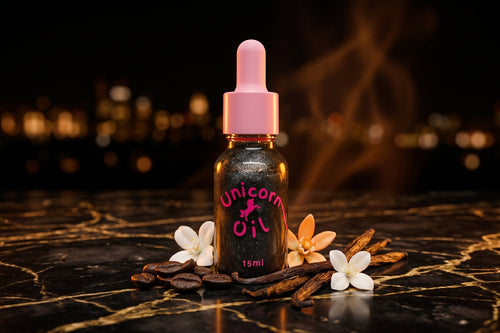 black shimmer Unicorn Oil bottle with pink dropper on black marble, styled with coffee beans, vanilla pods, white flowers, warm candlelight, and smoky midnight accents.

