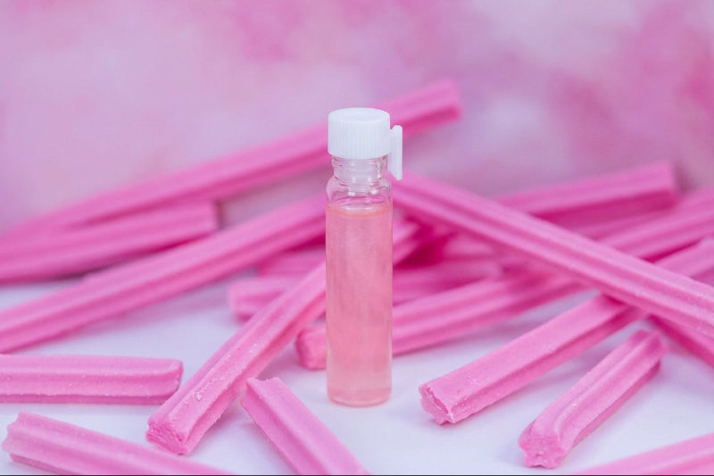 A small clear plastic bottle filled with pink liquid and topped with a white cap, standing upright on a white surface surrounded by scattered pink wax melts or candy sticks against a soft pink background.