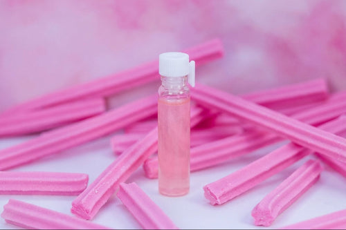 A small clear plastic bottle filled with pink liquid and topped with a white cap, standing upright on a white surface surrounded by scattered pink wax melts or candy sticks against a soft pink background.