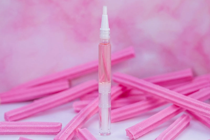 A clear cylindrical cosmetic vial with a white applicator cap stands upright on a white surface, surrounded by scattered pink textured sticks against a pink marbled background.