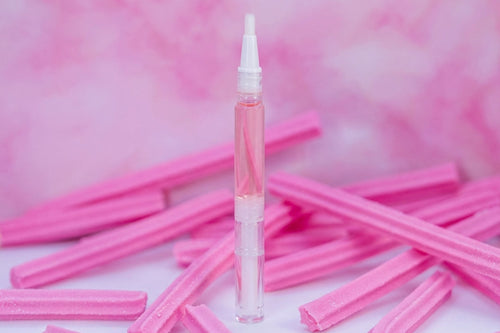 A clear cylindrical cosmetic vial with a white applicator cap stands upright on a white surface, surrounded by scattered pink textured sticks against a pink marbled background.