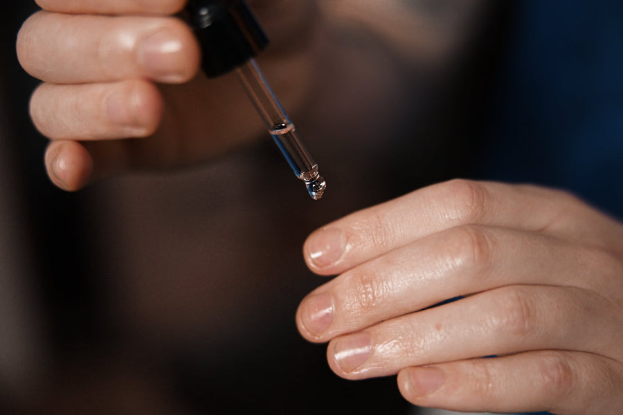 Close-up of a hand holding a dropper with a dark liquid, against a blurred background.