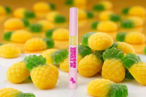 A pink lip balm tube labeled 'UNICORN CUTICLE OIL' stands upright in front of scattered pineapple-shaped gummy candies on a white surface with a yellow background.
