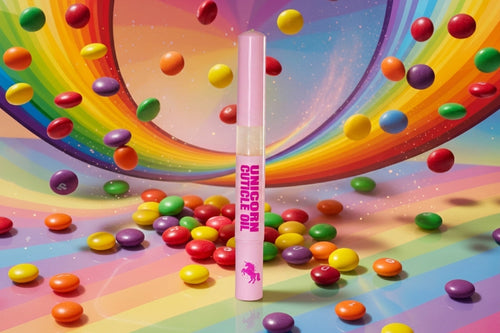 A pink tube of 'Unicorn Cuticle Oil' cosmetic product stands upright in the center of a vibrant, candy-themed scene filled with floating and scattered colorful candies on a rainbow-striped surface.
