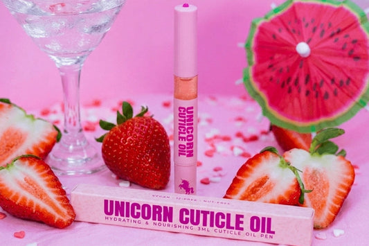 A pink cosmetic tube labeled 'UNICORN CUTICLE OIL' stands upright beside its matching pink box, styled with strawberries, sliced watermelon props, and a cocktail glass on a pink surface.
