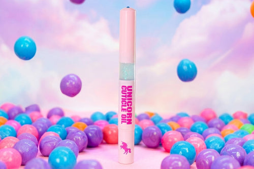 A pink cosmetic tube labeled 'UNICORN CUTICLE OIL' stands upright in a bed of pastel plastic balls, with a dreamy pink-and-blue cloud background and floating colorful spheres.