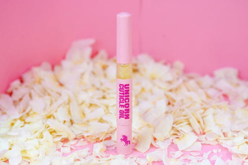 A pink tube of unicorn cuticle oil standing upright on a bed of pale flower petals against a solid pink background.