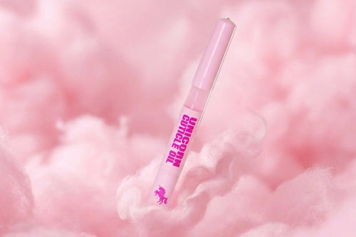 A pink cosmetic pen-style tube labeled 'UNICORN OIL' with a small unicorn logo, standing upright on fluffy pink cotton against a soft pink background.
