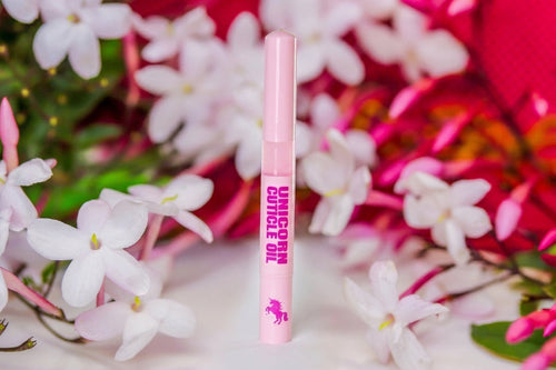 A pink cosmetic tube labeled 'UNICORN OIL stands upright against a soft-focus floral background of white blossoms and bright pink flowers.