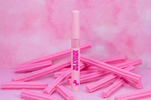 A pink tube labeled 'UNICORN OIL' stands upright in the center, surrounded by scattered pink candy sticks on a pink marbled background.