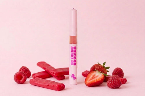 A tube of unicorn cuticle oil stands upright in the center, surrounded by red raspberry and strawberry garnishes and pink-red wax melts on a soft pink background.