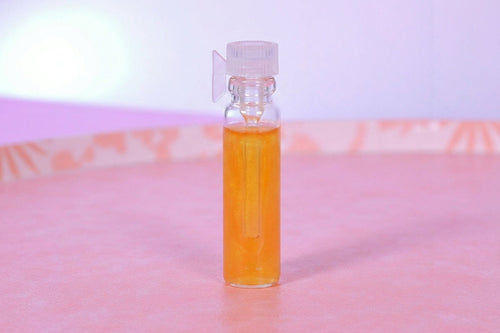 A small clear glass vial filled with bright orange liquid, topped with a translucent cap and a small clear plastic clip, standing on a pink surface against a soft pink and purple background.