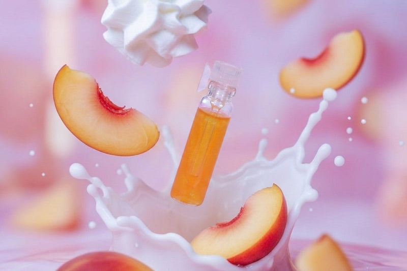 A small clear bottle filled with orange liquid floating in a pink studio scene with peach slices and a white splash around it.