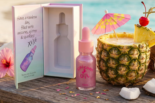 Unicorn oil bottle with dropper, gift box, pineapple with candle, and flowers on a wooden surface.