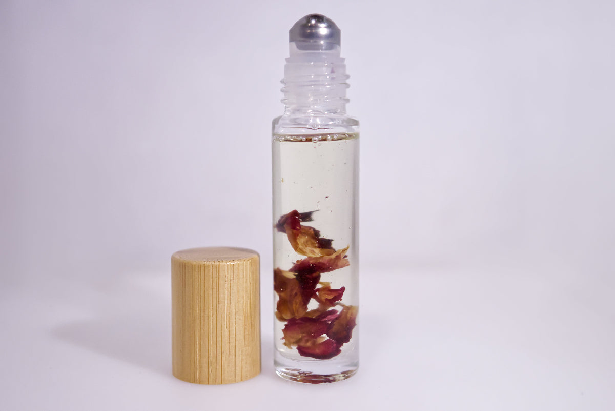 A clear glass roller bottle filled with clear liquid and dried red flower petals, shown next to its light wood cap on a plain light gray background.