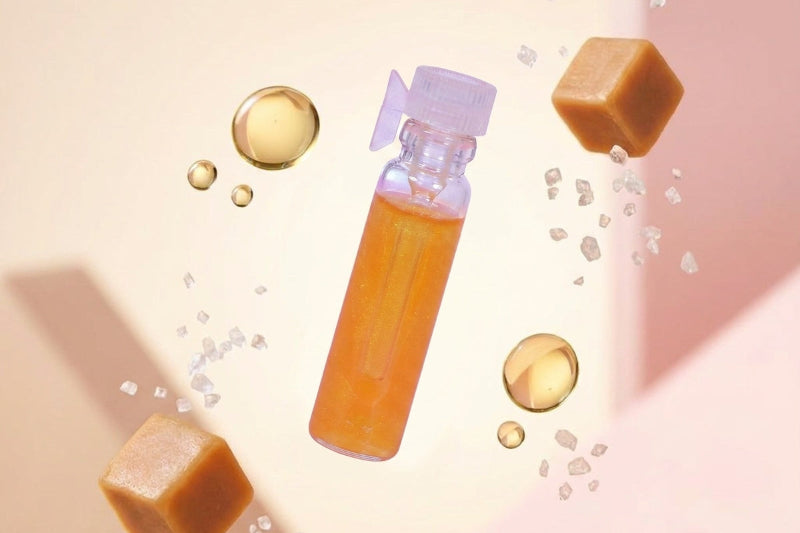 A clear plastic bottle filled with amber-orange liquid and topped with a translucent purple cap, floating among caramel cubes, gold droplets, and sugar crystals on a soft peach background.
