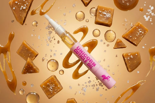 A cosmetic dropper bottle labeled 'UNICORN CUTICLE OIL' in pink and white, surrounded by floating caramel pieces, caramel drizzle, and clear bubbles on a warm beige background.