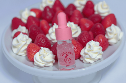 5ml glass dropper bottle of Strawberries & Cream Unicorn Cuticle Oil featuring pink shimmering oil and a professional boutique label surrounded by a plate of strawberries