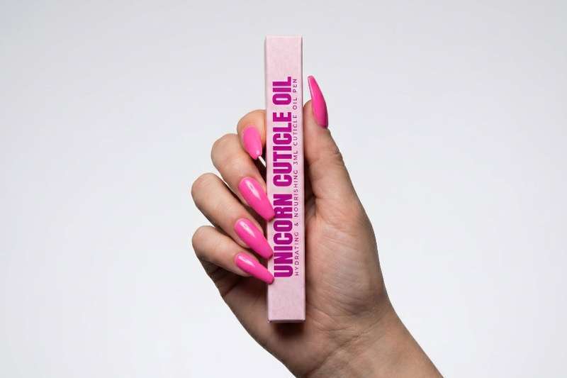 Hand holding a pink tube of 'Unicorn Cuticle Oil' with pink nail polish on a light gray background