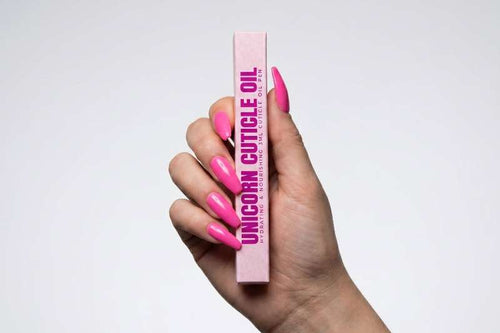 Hand holding a pink tube of 'Unicorn Cuticle Oil' with pink nail polish on a light gray background