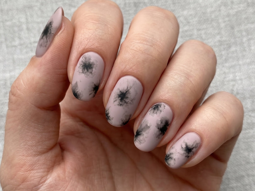 Hand with pink nails featuring black floral patterns on a light gray background