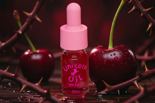 A 5ml dropper bottle of pink cherry-flavored 'Unicorn Oil Mini' centered between two fresh, glistening red cherries and dark thorny branches on a dark reflective surface.
